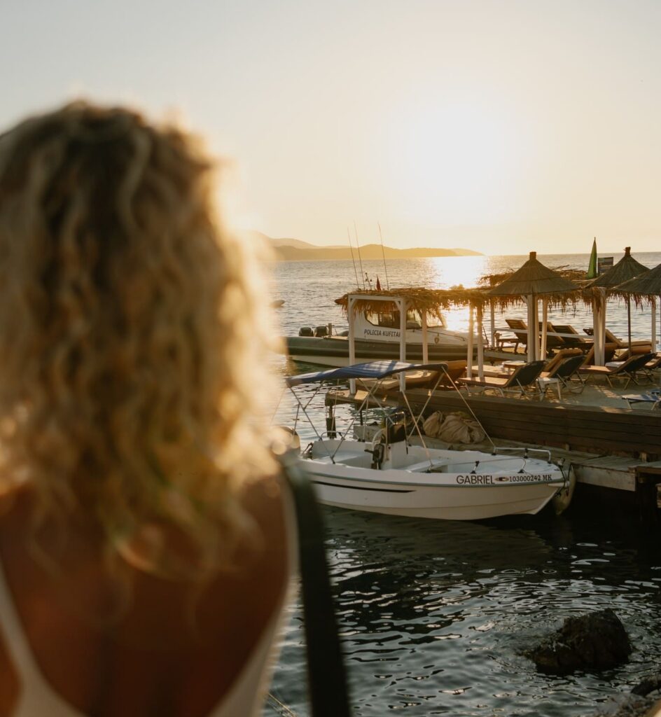 blonde woman watching sunset over boats and beach huts in Albania